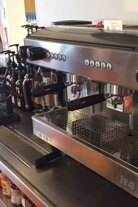 the inside of a coffee shop you see an espresso grinder next to several bottles of coffee syrup lined up next to a new two group commercial espresso machine with two steam wands