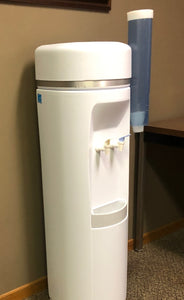 a white office water cooler shown in a professional office breakroom with a cup dispenser on the right side filled with cone water cups and a water filter system in the back of the cooler