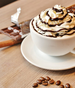 Mocha latte with Hollander chocolate drizzled on top of white whipped cream in a white coffee cup with a few whole coffee beans spread throughout the foreground of this coffee shop counter 