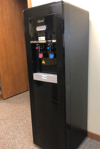 this black high end water cooler has a red hot water faucet and a cold blue colored faucet with a grey drip tray in the hallway of a commercial building using reverse osmosis to treat the water
