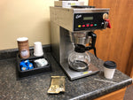 a Curtis coffee brewer with cups, lids, creamer and sugar set up next to it in an office setting