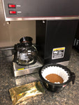 a Bunn commercial coffee brewer with a glass pot on the burner a gold foil packet of coffee in the foreground and to the right a black brew basket filled with light brown ground coffee